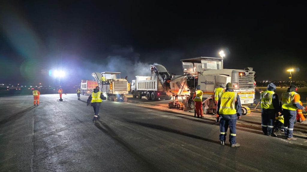 Three Injumo Development workers in safety gear hand-painting precise yellow markings on an airport apron at night. They are illuminated by work lights and vehicle headlights, with parked airplanes in the background.