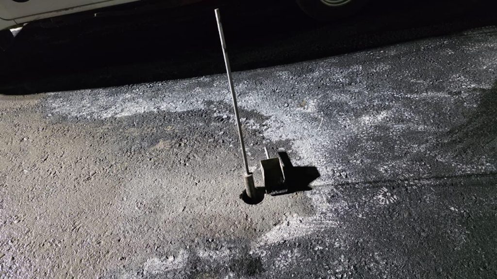 Close-up, high-angle view of a circular core hole drilled into asphalt pavement. A metal measuring rod is inserted vertically into the hole, casting a sharp shadow. White spray paint markings surround the area on the textured black tarmac.