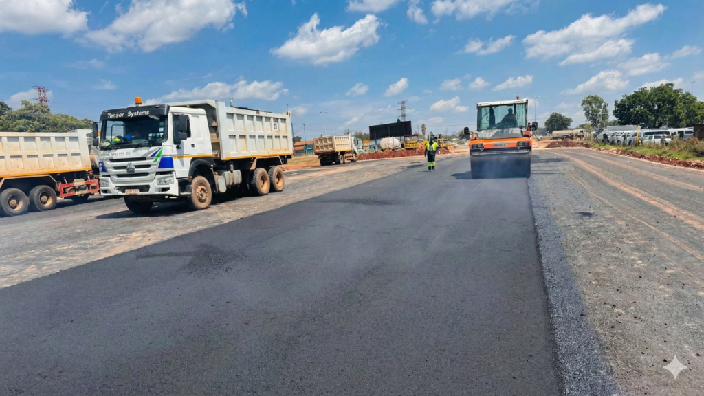 A wide shot of a road construction site featuring a white dump truck on the left and an orange road roller compacting fresh asphalt on the right, set against a bright blue sky with scattered clouds.