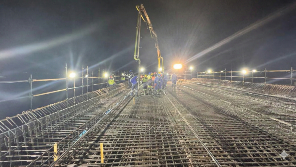 Nighttime construction site view for Injumo Development, showing a dense steel rebar grid, bright floodlights, and a crew of workers using a concrete pump boom to pour a large deck.