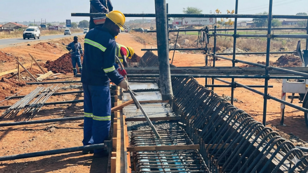 Construction worker wearing a yellow hard hat and safety gear guiding a concrete pump hose into a steel-reinforced trench.