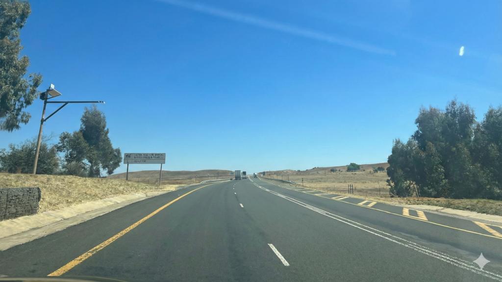 A view from a vehicle driving down a paved two-lane highway under a clear blue sky, flanked by dry grassy fields and trees, with a roadside sign displaying 'Routine Road Maintenance' information.