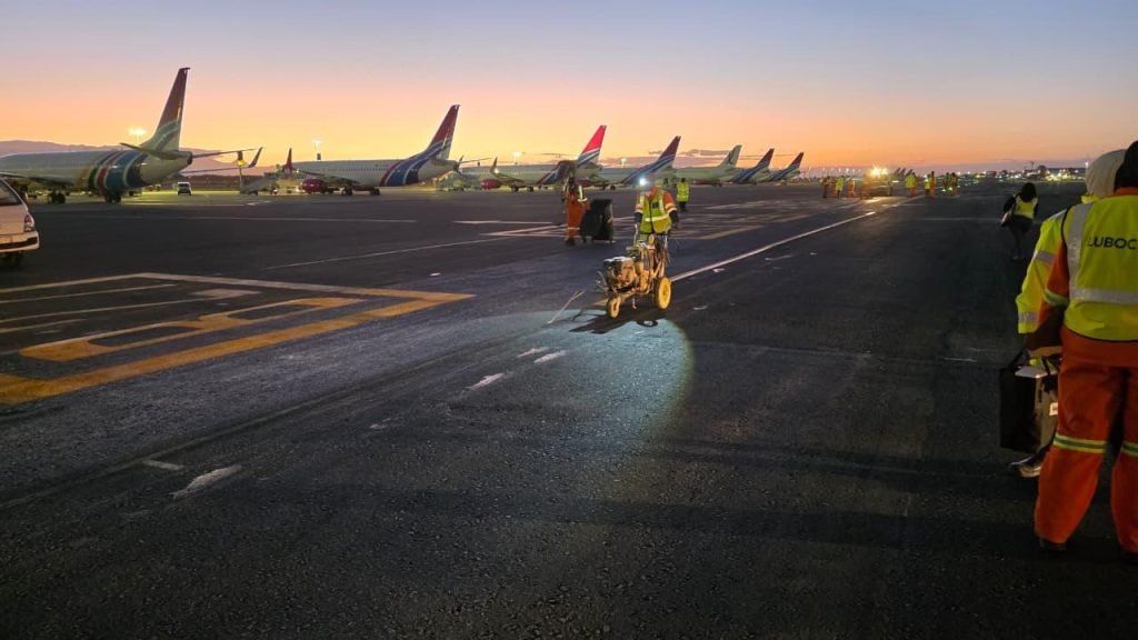 Wide shot of an airport apron at twilight with a vibrant orange and purple sky. In the foreground, Injumo Development workers in high-visibility vests operate a line-marking machine to paint fresh white lines on the asphalt. In the background, a row of Airlink Embraer jets are parked, facing left.
