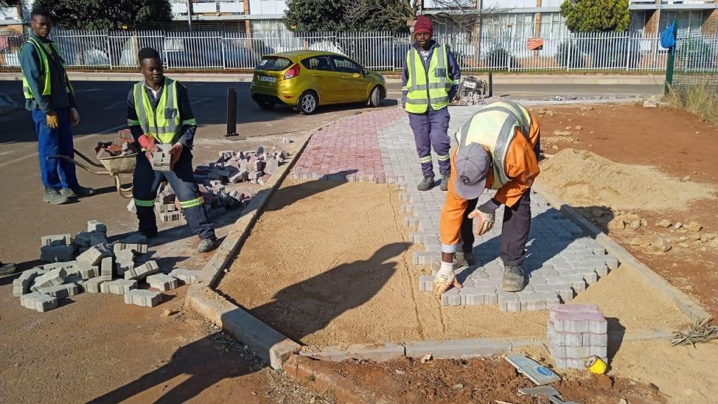 A construction crew from Injumo Development installing a new paved sidewalk. In the foreground, a worker in an orange shirt bends down to precisely place grey interlocking bricks onto a sand bed. Behind him, another worker carries a stack of pavers, while two colleagues in high-visibility yellow vests supervise the site. A yellow car and a white apartment complex fence are visible in the background.